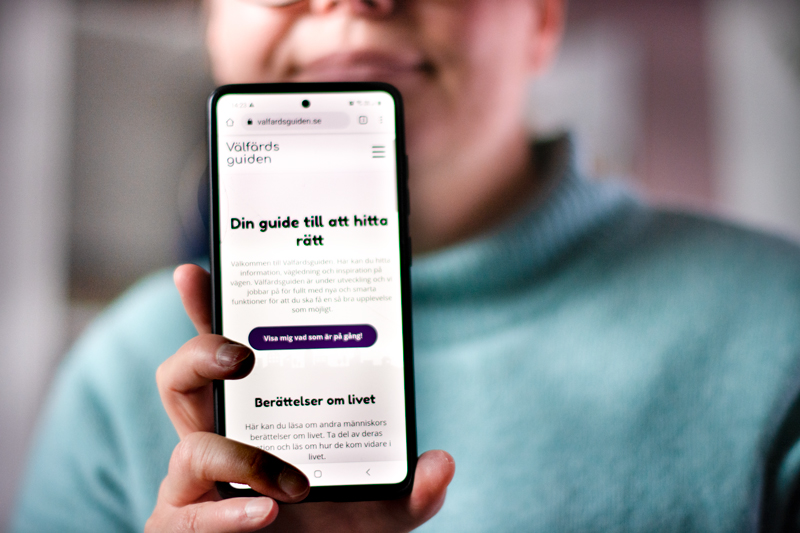 En person håller upp en mobiltelefon med Välfärdsguiden på skärmen.